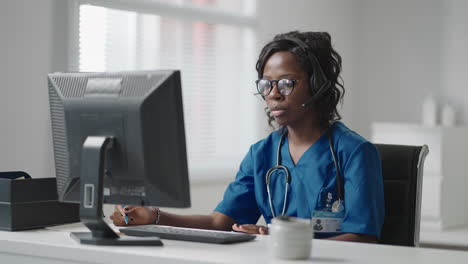 African-american-black-female-general-practitioner-in-white-coat-sitting-at-desk-in-doctor's-office-and-scrolling-computer-mouse-while-reading-patient's-medical-history